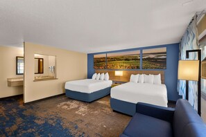 A hotel room with two beds, a large window with a landscape view, a bathroom with a mirror and sink, and a blue carpet.
