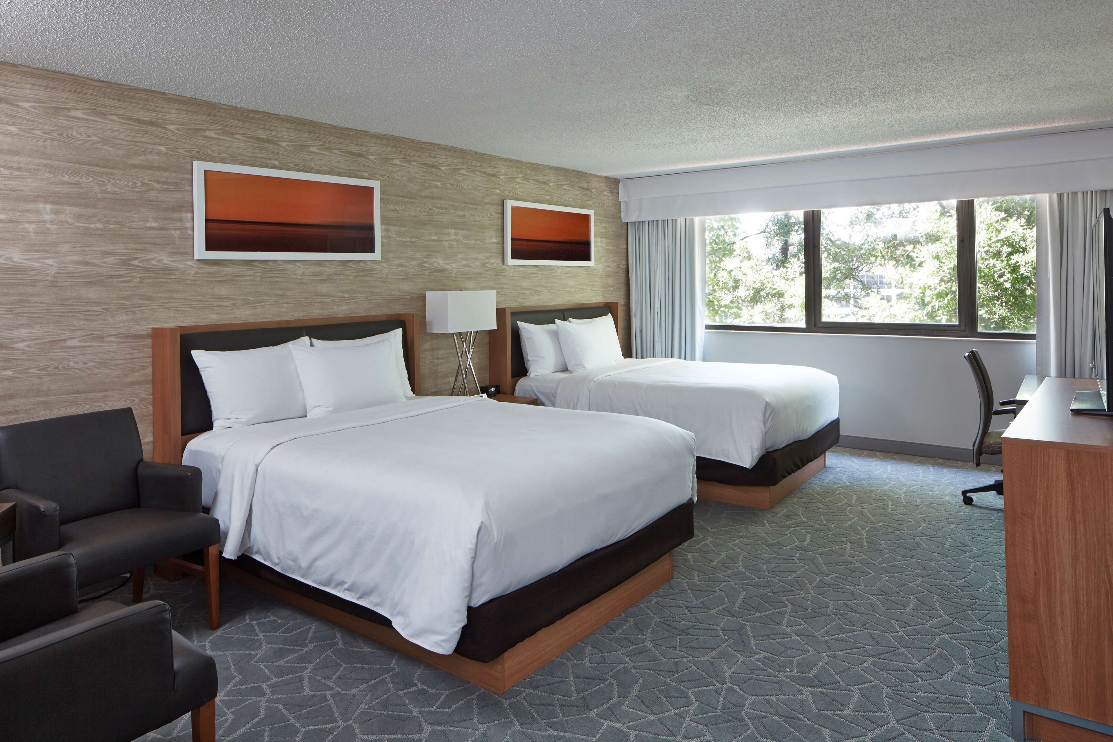 A hotel room with two beds, a desk, a chair, and a window with a view of trees.