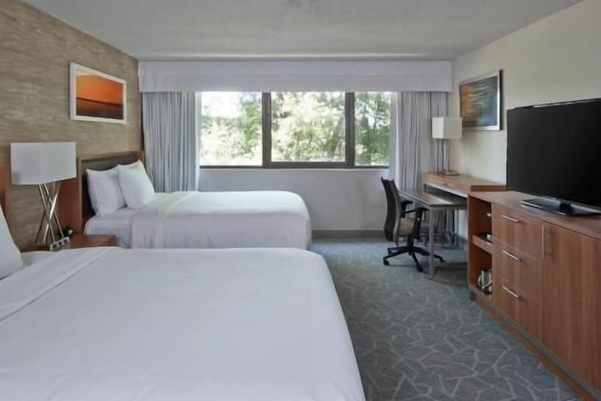 A hotel room with two beds, a desk, a television, and a window with a view of trees.
