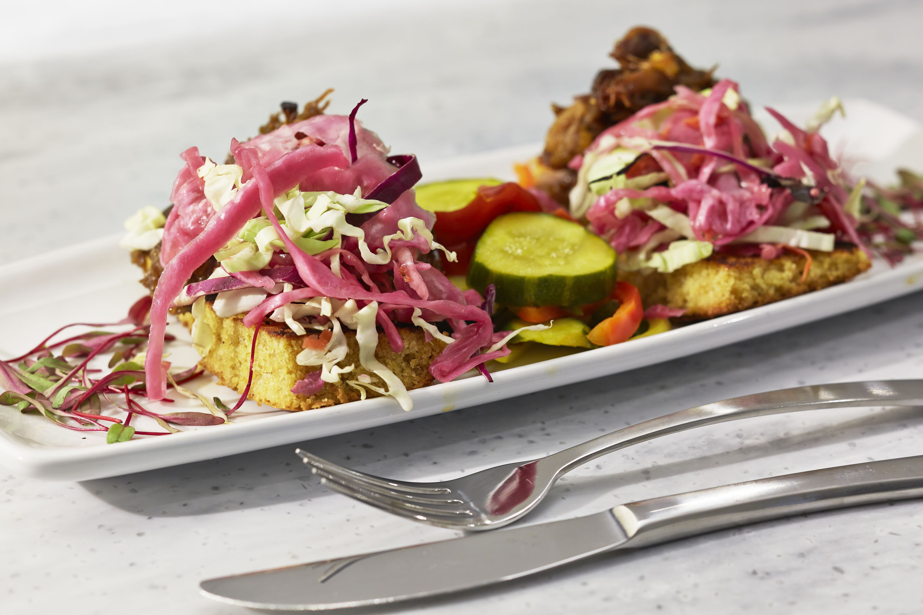 A white rectangular plate with a sandwich topped with red cabbage and sprouts, a slice of cucumber, and a piece of grilled meat.