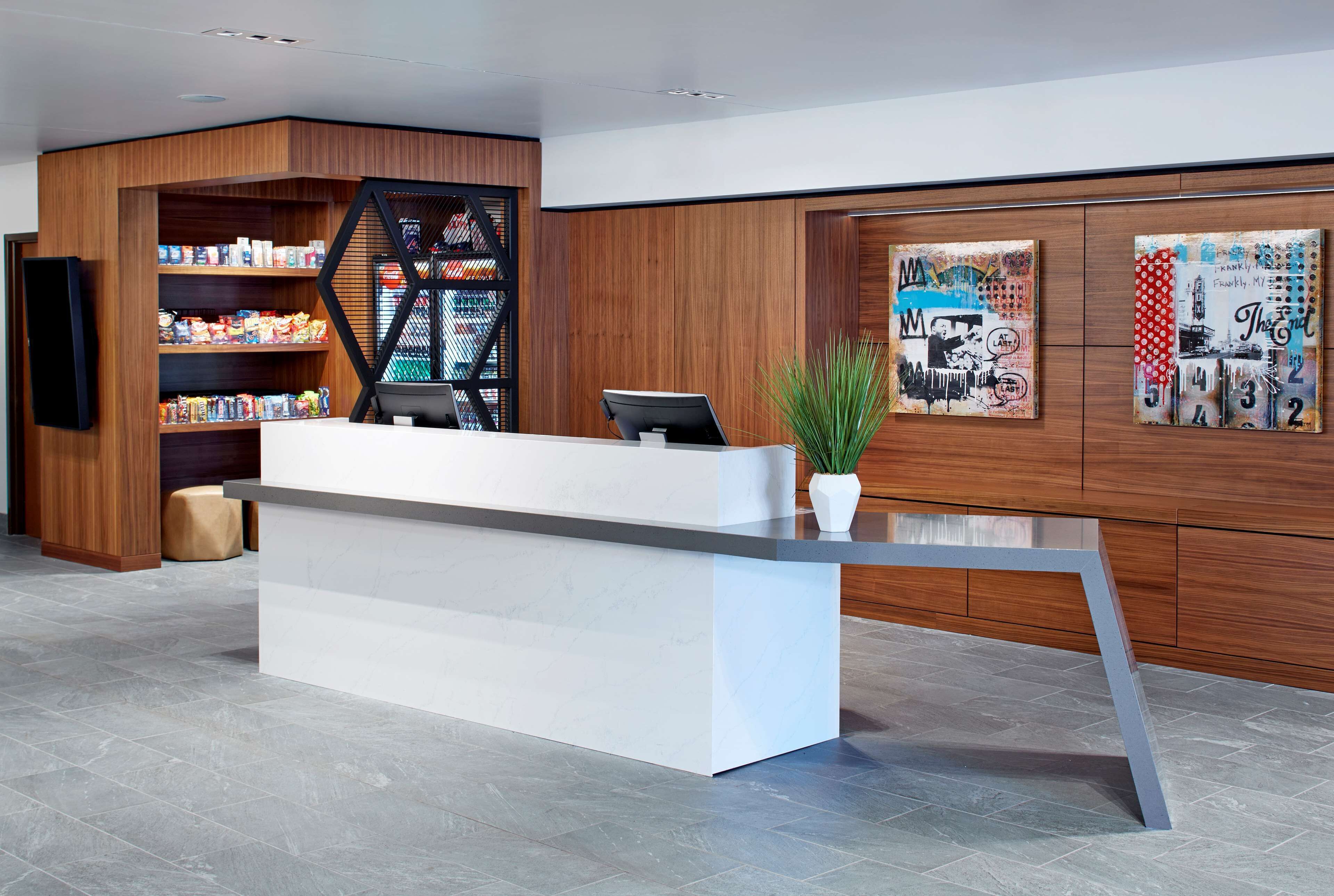 A modern reception desk with a white counter and a computer monitor. To the left, there is a wooden shelving unit with various items and a black geometric partition. On the right, there are two pieces of wall art depicting urban scenes.