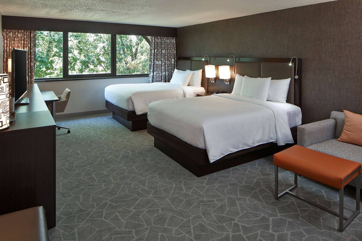 A hotel room with two beds, a desk with a chair, a sofa, and a bench.
