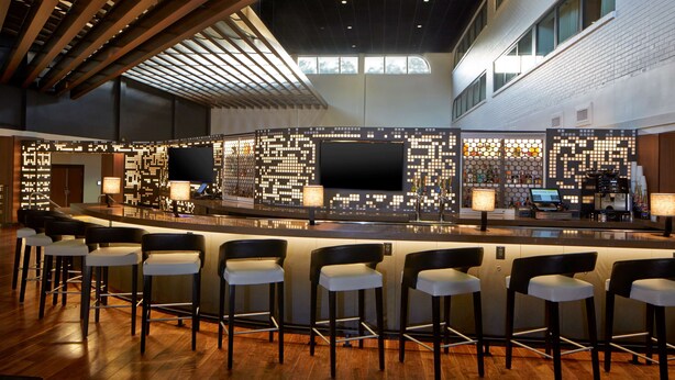 A modern bar area with a long counter, multiple bar stools, and a high ceiling with exposed beams. - DoubleTree by Hilton Atlanta Perimeter Dunwoody (Atlanta)