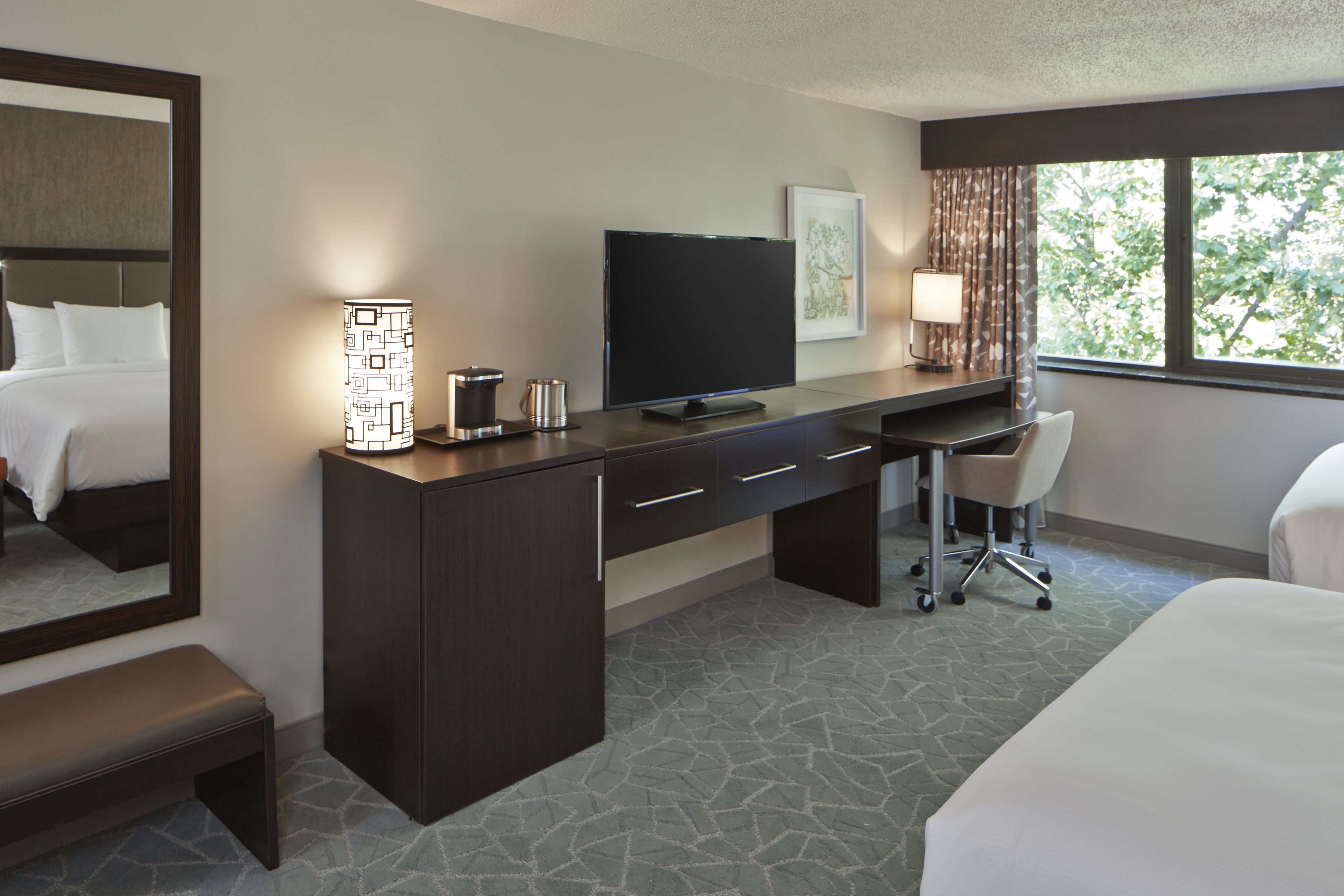 A hotel room with a bed, a desk with a flat-screen TV, a chair, a lamp, and a large window with a view of trees.