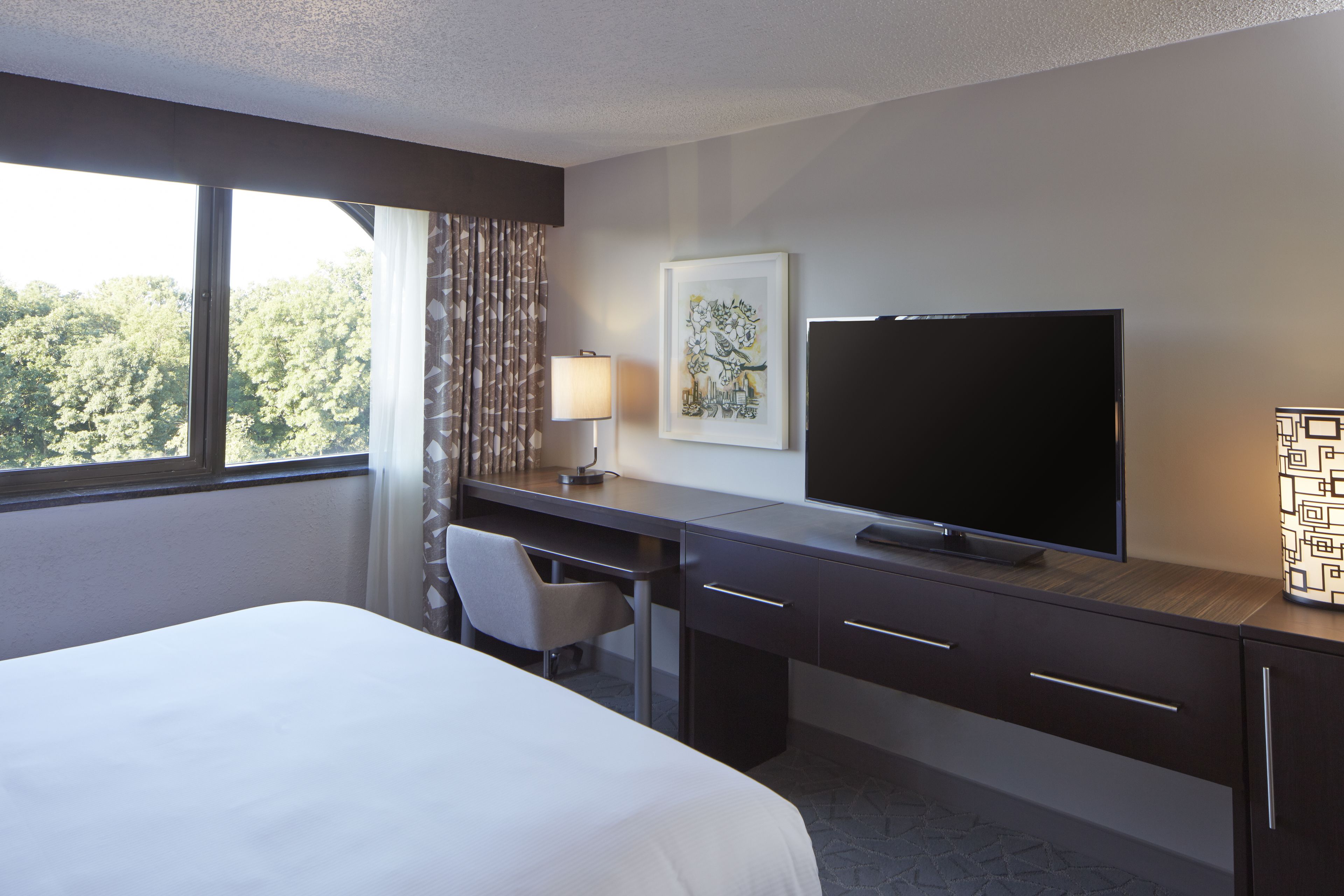 Une chambre d’hôtel avec un lit, un bureau, une chaise, une télévision et une fenêtre avec des rideaux.
