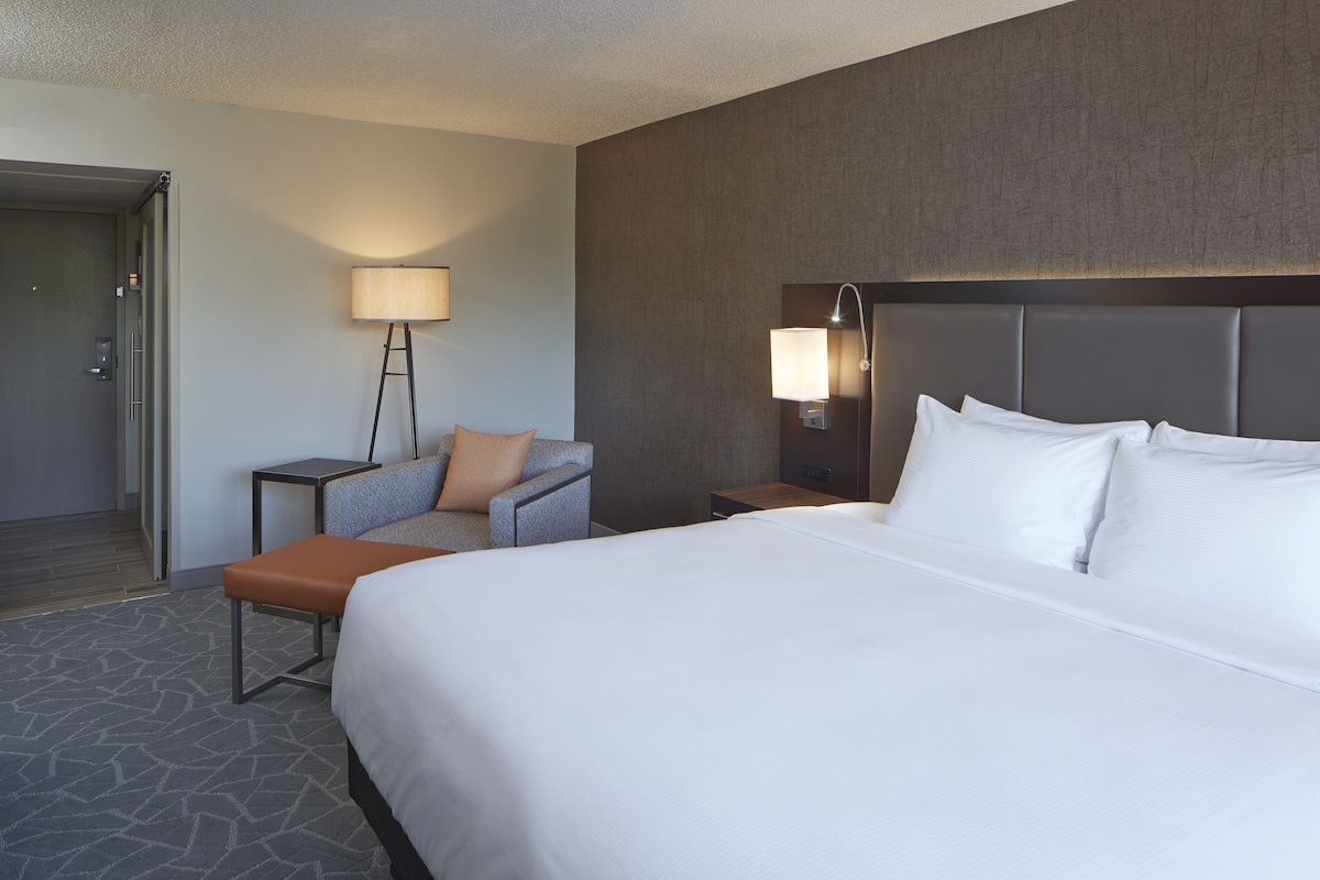 Ein Hotelzimmer mit einem großen Bett, einem Sessel, einem Nachttisch, einer Lampe und einer Tür.