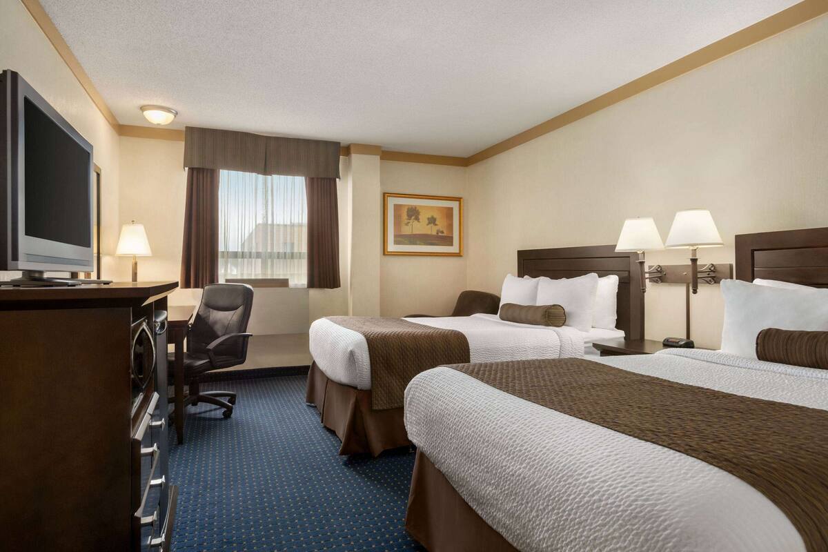 Ein Hotelzimmer mit zwei Betten, einem Schreibtisch mit Fernseher, einem Sessel und einem Fenster mit Vorhängen.