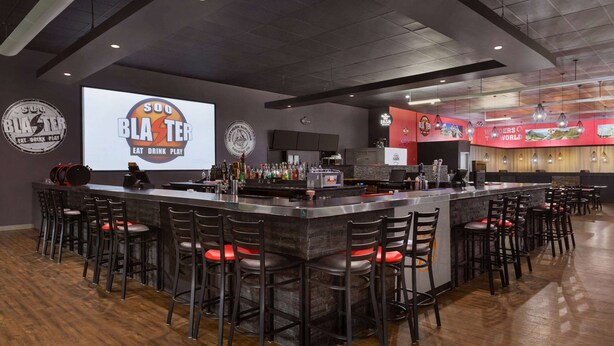 A bar area with a large screen displaying 'Soo Blaster' branding, bar stools, and a counter with various bottles. - Days Inn & Suites by Wyndham Sault Ste. Marie ON (Sault Ste. Marie)
