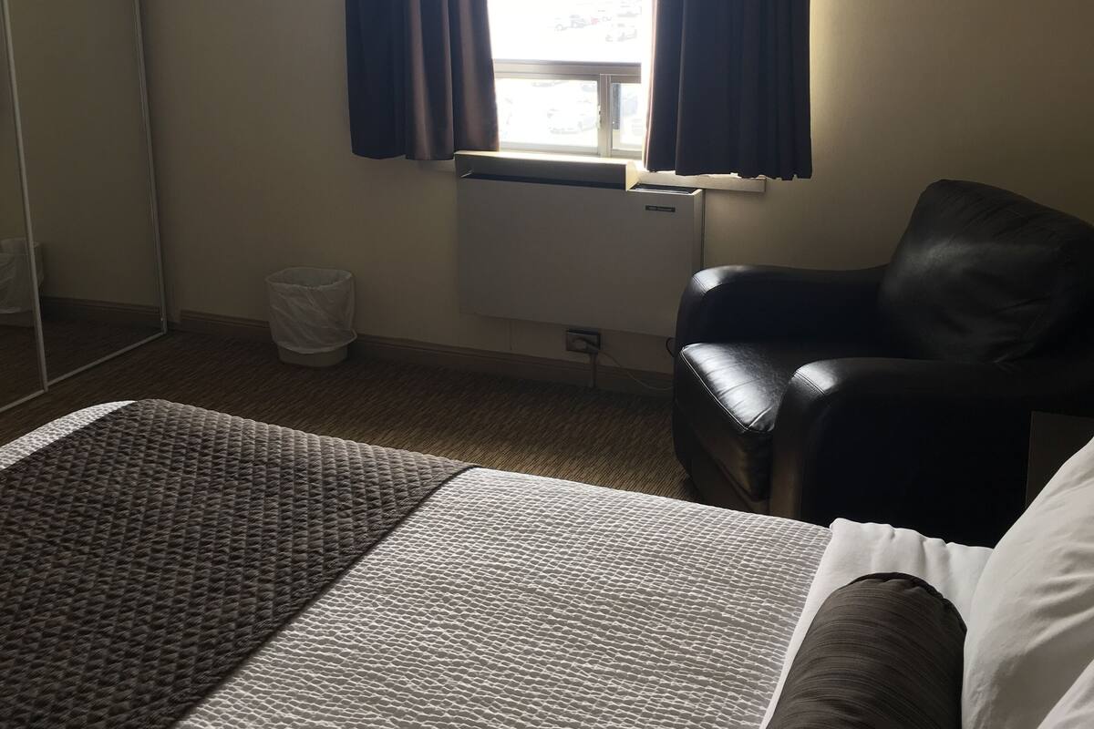 Ein Hotelzimmer mit Bett, Sessel, Fenster mit Vorhängen, Heizkörper und Mülleimer.