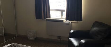 Habitación de hotel con cama, silla, ventana con cortinas, radiador y un cesto de basura.