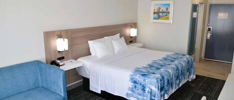 A hotel room with a bed, a blue armchair, a bedside table, a nightstand, a wall-mounted lamp, a painting, and a door.