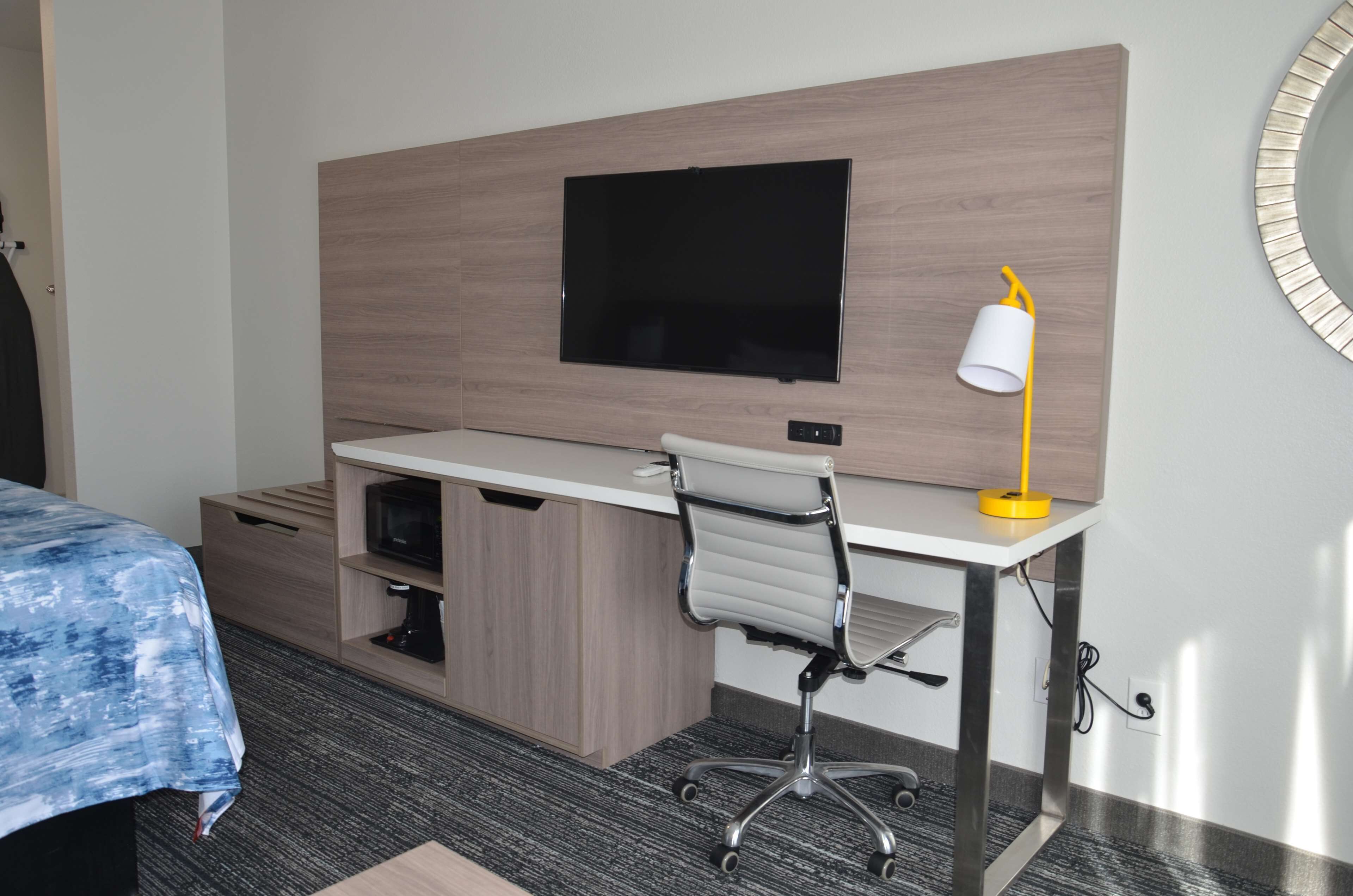 A hotel room with a bed, a desk with a chair, a TV mounted on the wall, and a small table with a lamp.