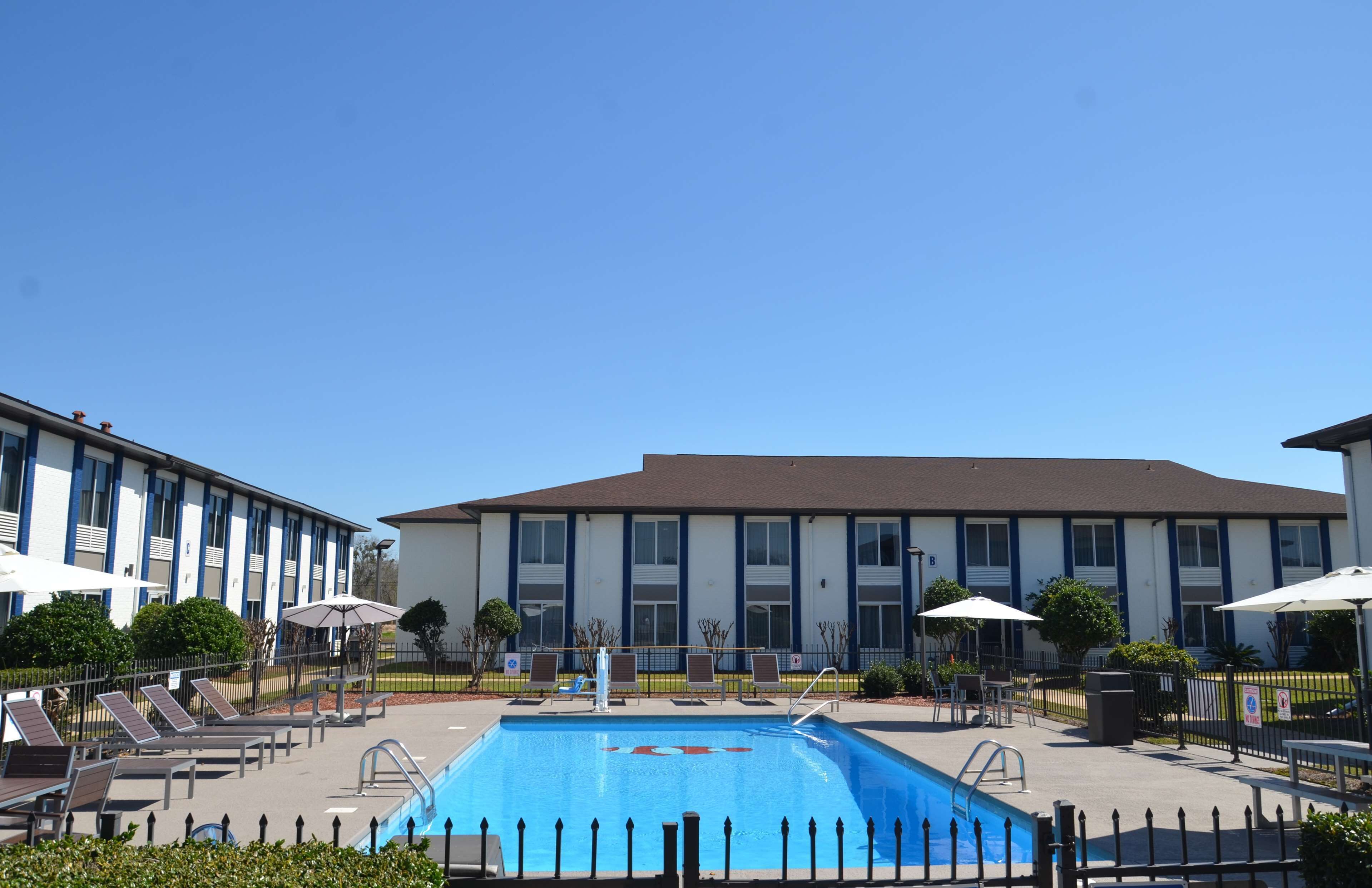 A swimming pool area with lounge chairs and umbrellas, surrounded by a fence, with a building in the background.