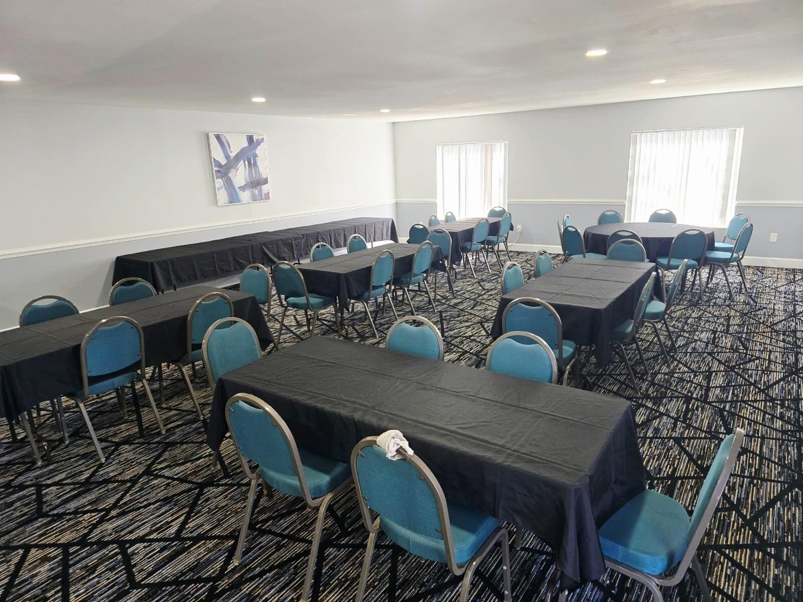 Un salón de conferencias con varias mesas y sillas, suelo alfombrado y amplias ventanas con persianas.