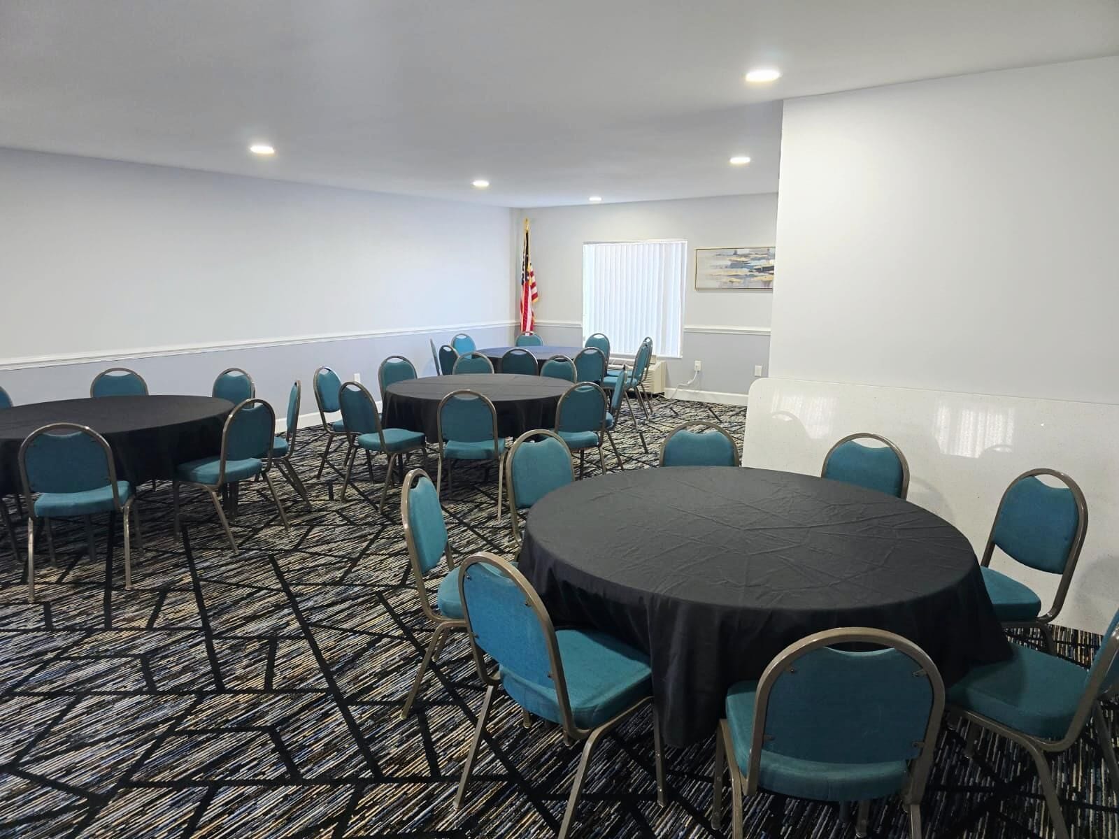 Un salón de conferencias con mesas redondas y sillas azules, suelo alfombrado y una bandera en la pared.