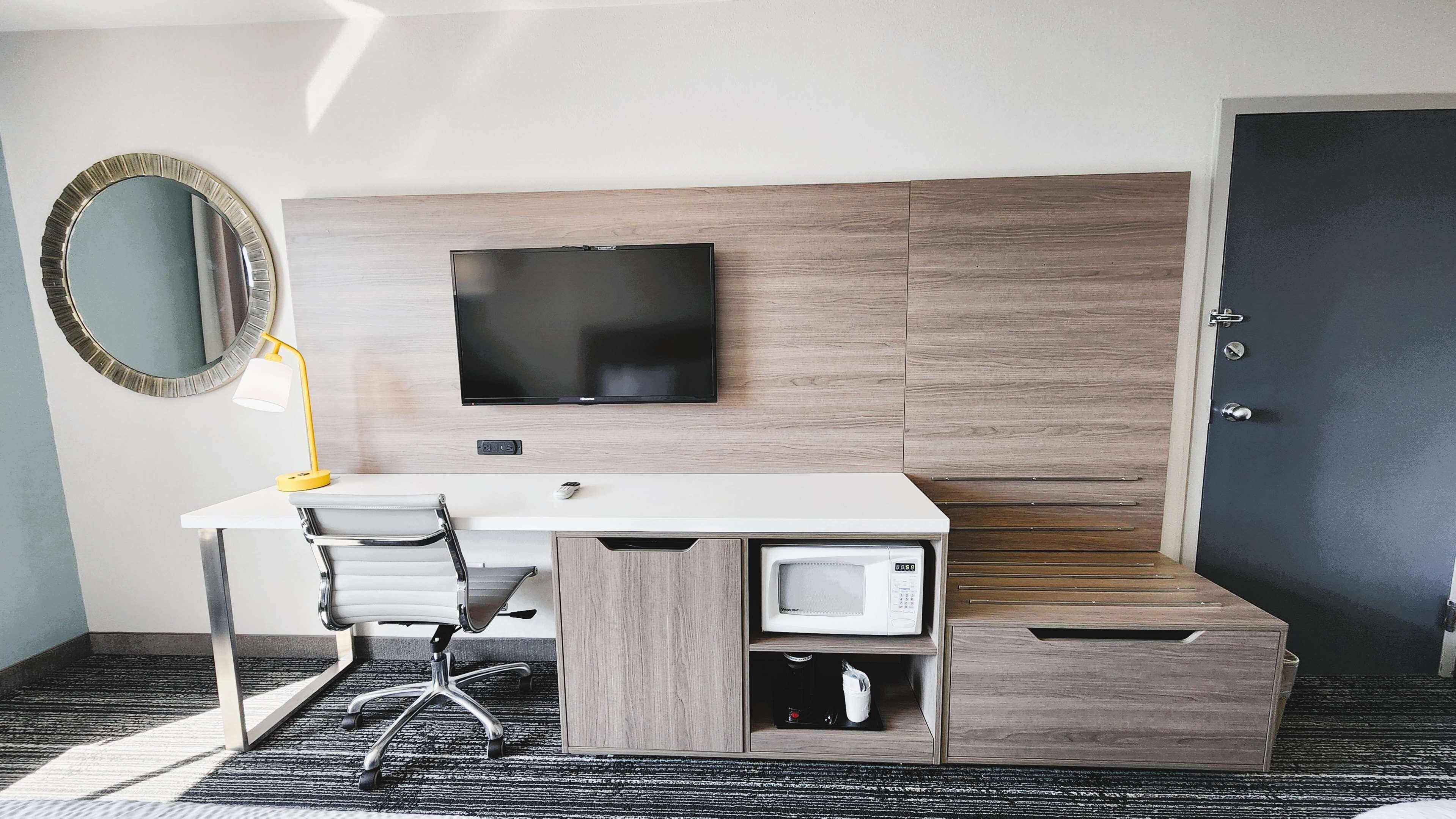 A hotel room with a desk, chair, TV, microwave, and a door.