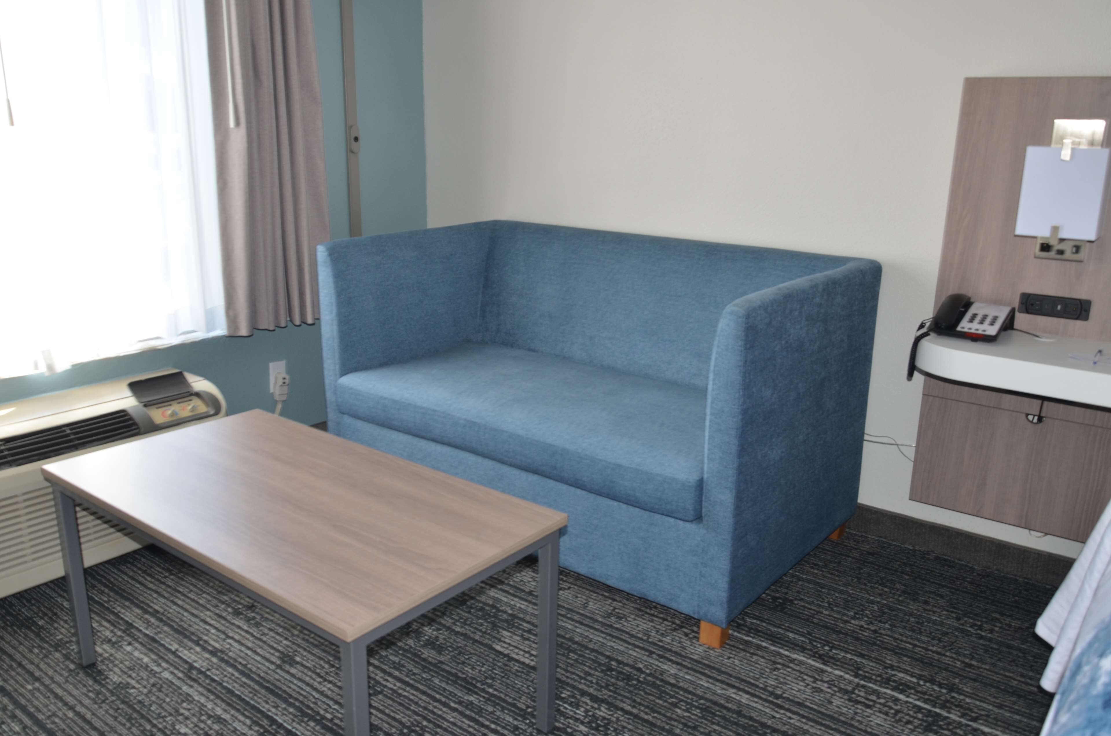 A hotel room with a blue sofa, a small wooden table, a desk with a telephone, and a mirror on the wall.