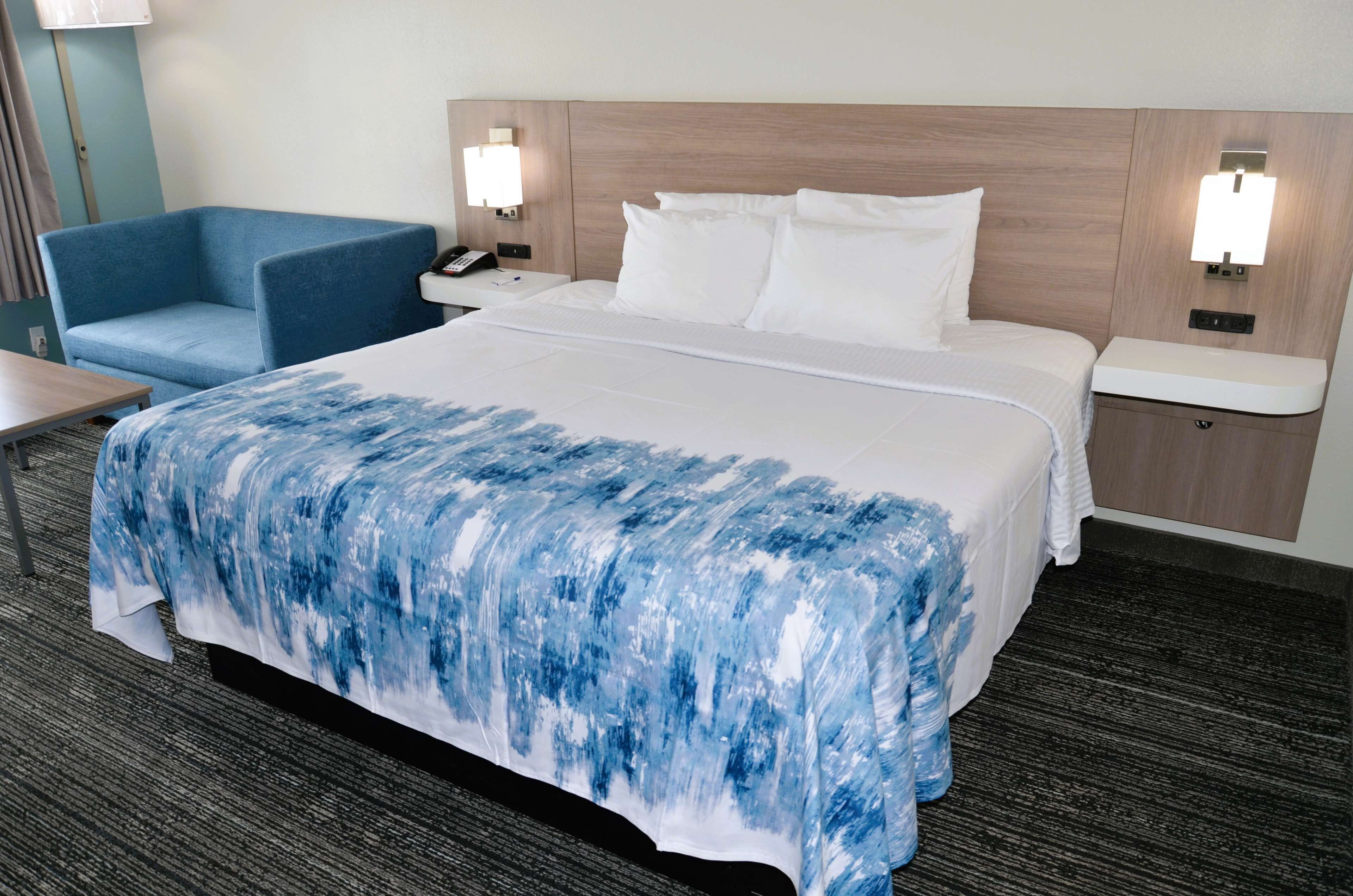 A hotel room with a large bed, a blue armchair, bedside tables with lamps, and a carpeted floor.