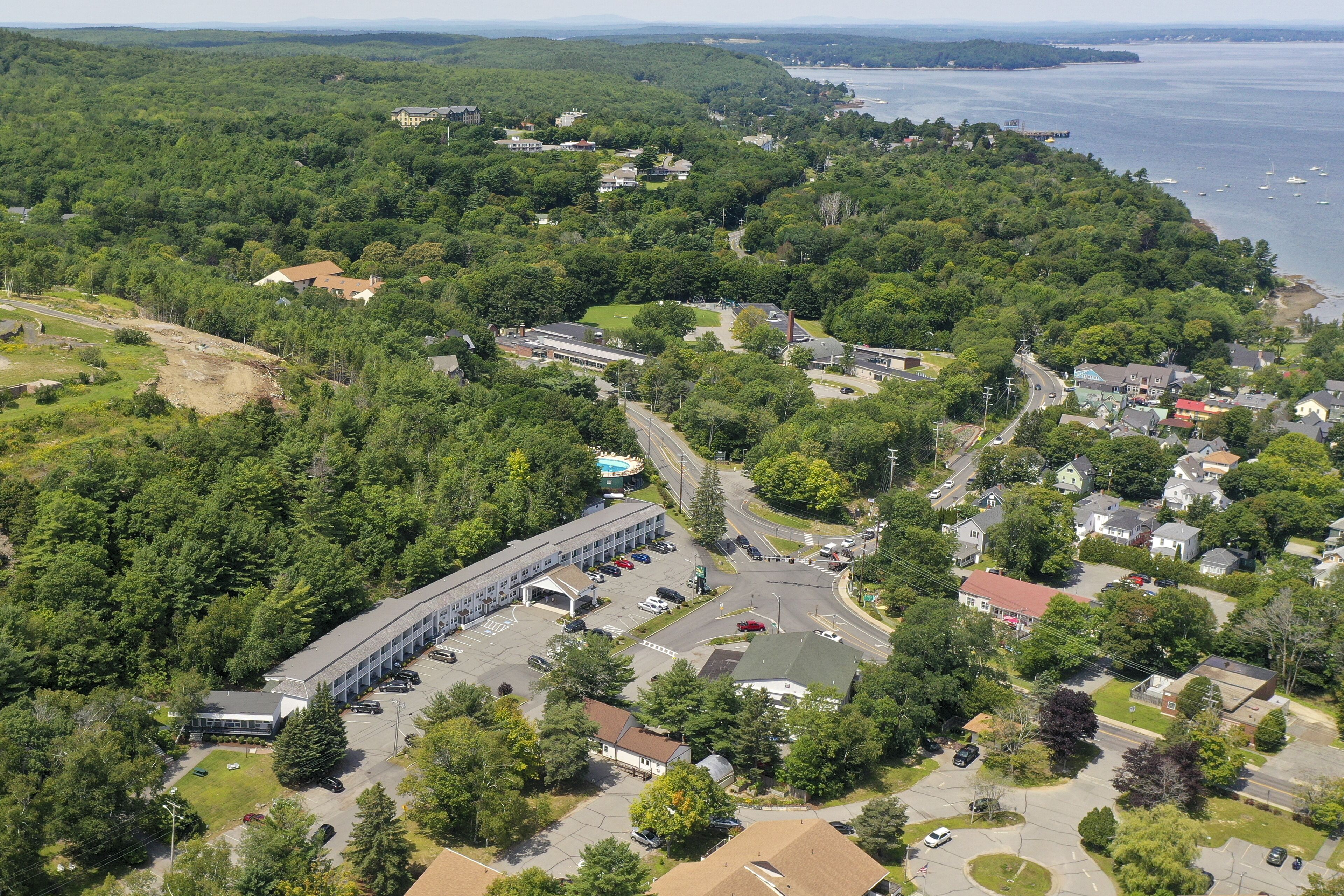 An aerial view of a residential area with a large building complex, surrounded by dense greenery and a coastline.