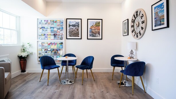 A modern room with a wooden floor, a round table, and four blue chairs. There are framed pictures on the wall and a vase with flowers on the windowsill.
