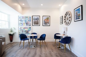A modern room with a wooden floor, a round table, and four blue chairs. There are framed pictures on the wall and a vase with flowers on the windowsill.