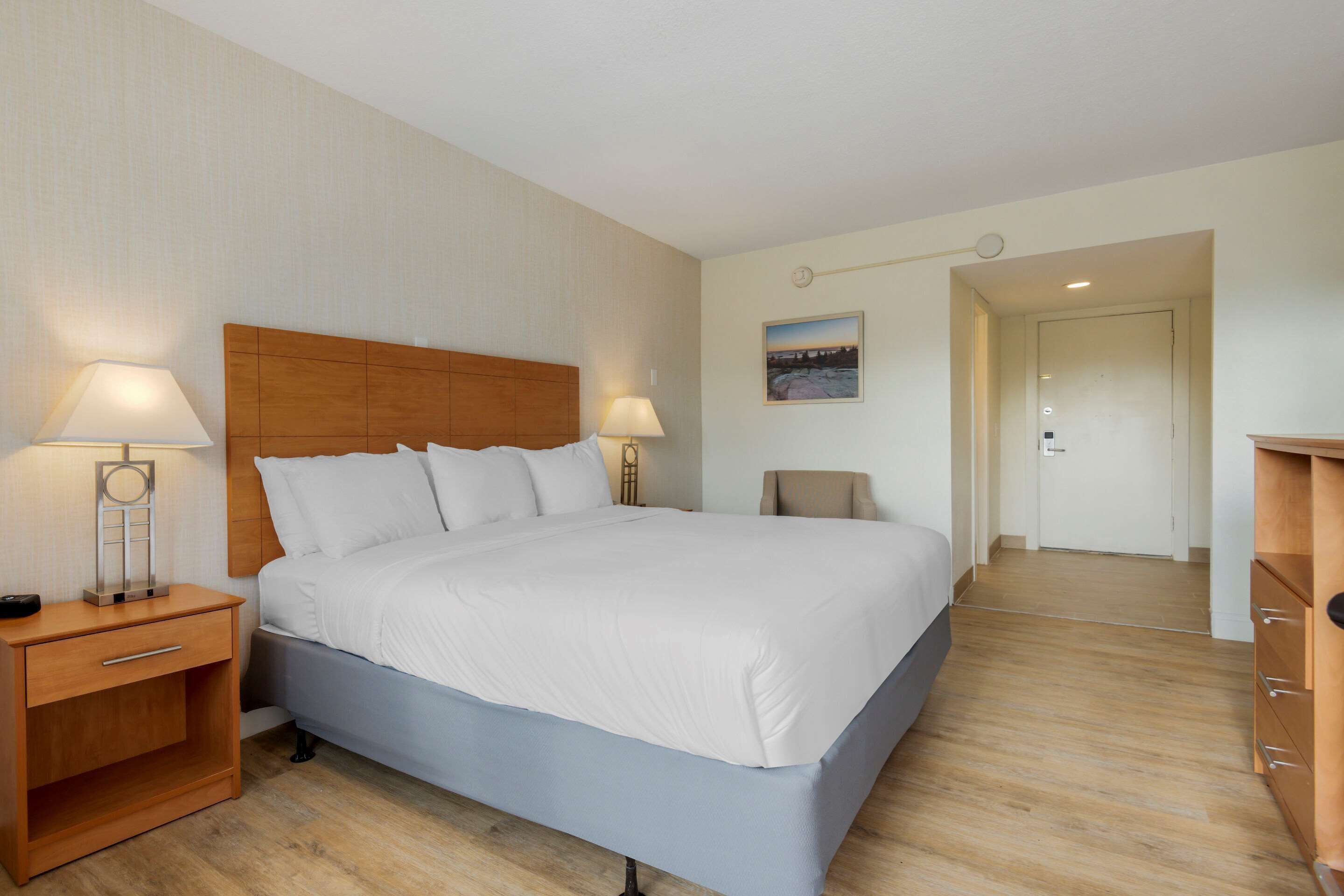 Una habitación de hotel con una cama grande, cabecera de madera, dos mesitas de noche con lámparas, una silla, un cuadro en la pared y una puerta que da a otra habitación.