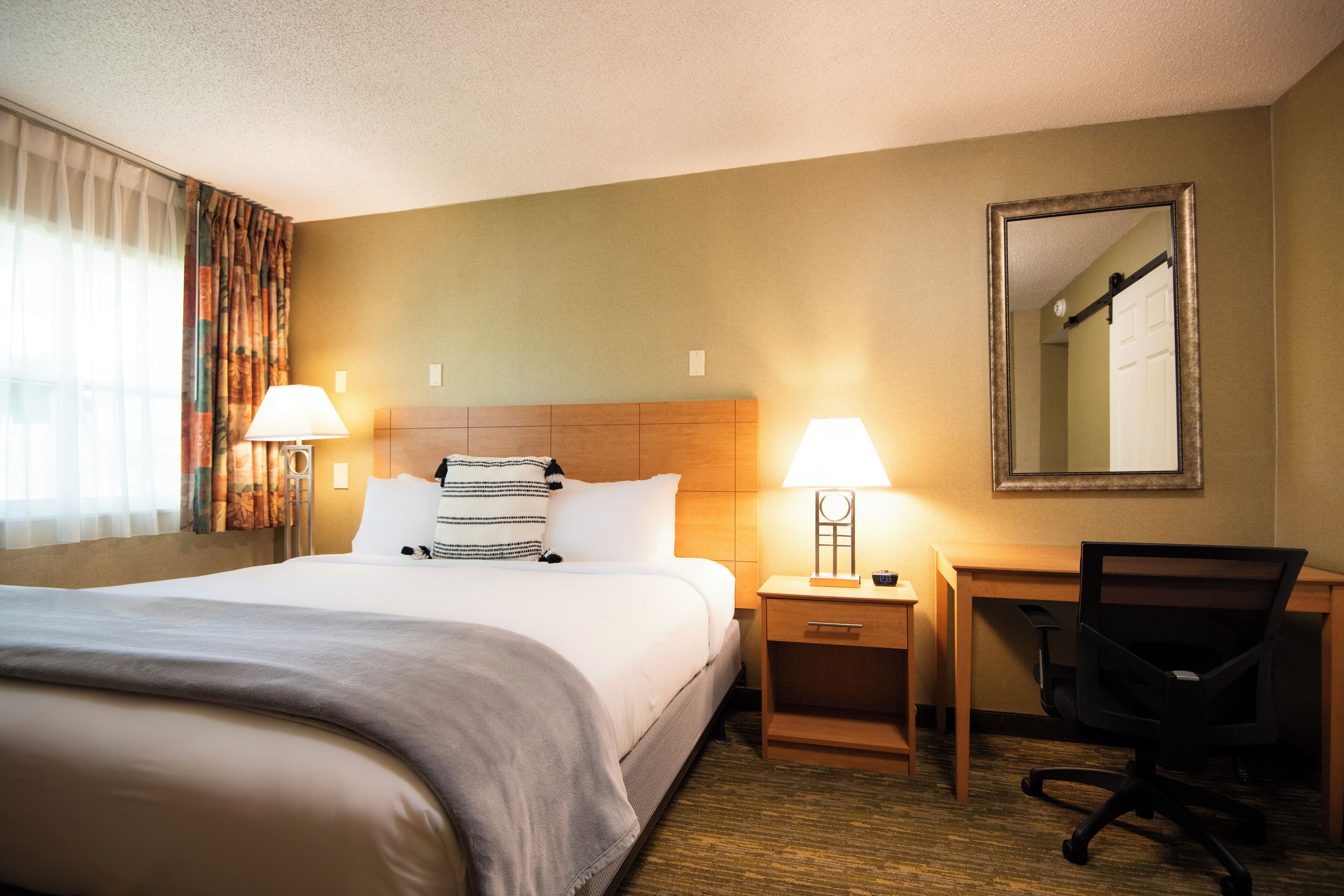 A hotel room with a bed, bedside lamps, a desk with a chair, and a mirror on the wall.