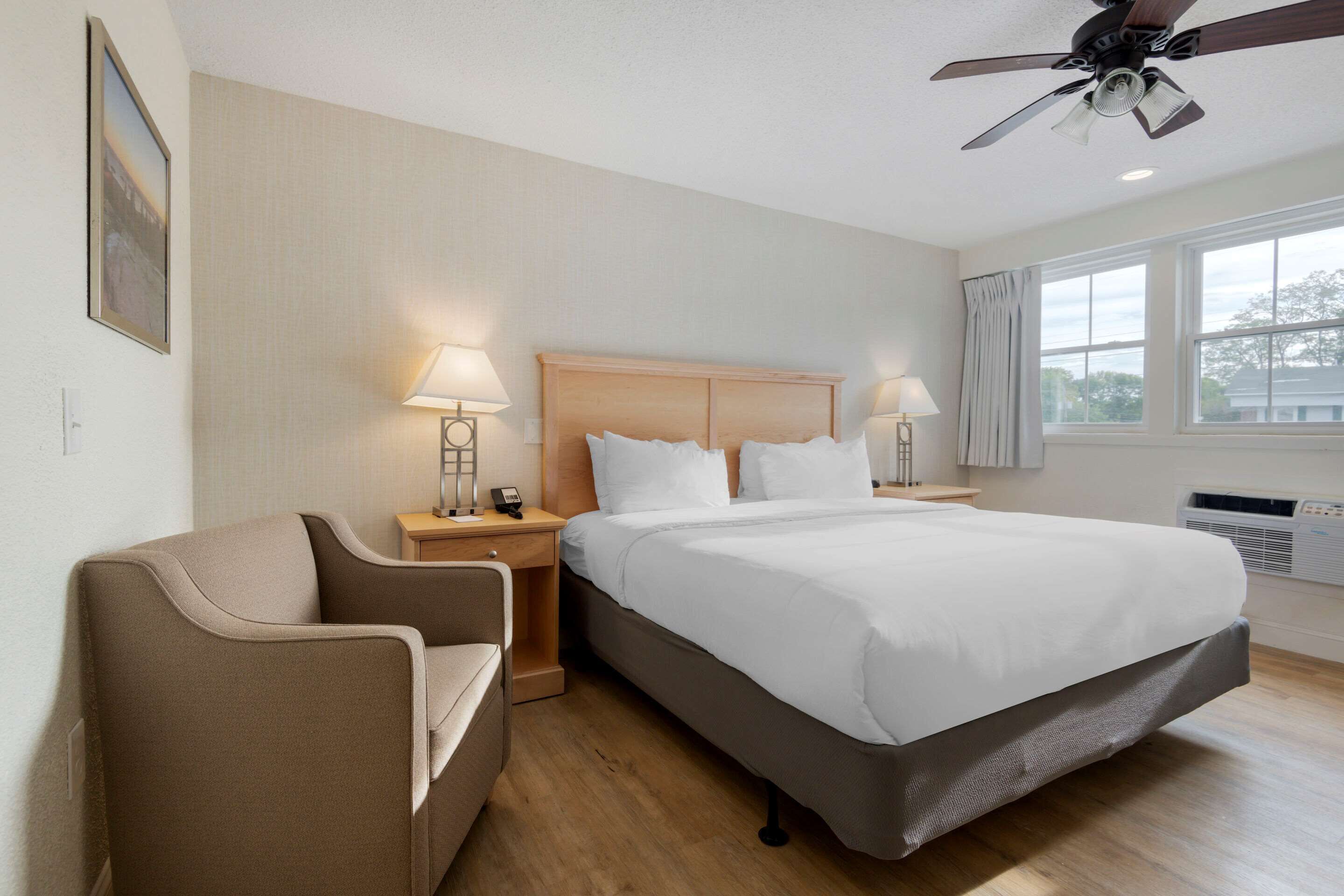 Une chambre d’hôtel comprenant un lit, une chaise, deux tables de chevet avec des lampes, un ventilateur de plafond et une fenêtre avec des rideaux.