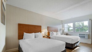 A hotel room with two beds, a wooden headboard, a nightstand with a lamp, and a large window offering a view of trees.