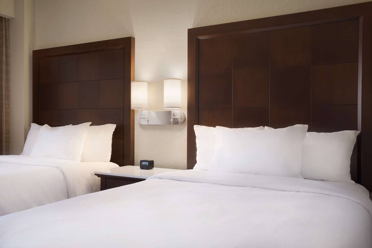A hotel room with two beds, a nightstand, and a wall-mounted lamp.