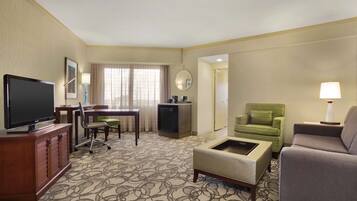A hotel room with a television, a desk, a green armchair, a grey sofa, and a patterned carpet.