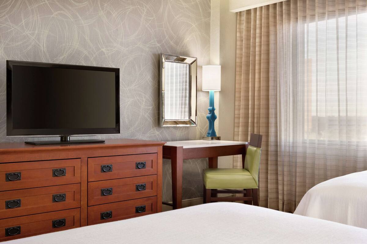 A hotel room with a TV on a wooden dresser, a desk with a chair, and a bed.
