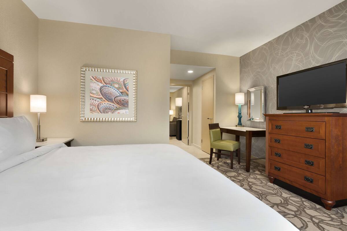 A hotel room with a large bed, a desk with a chair, a dresser, a television, and a decorative wall piece.