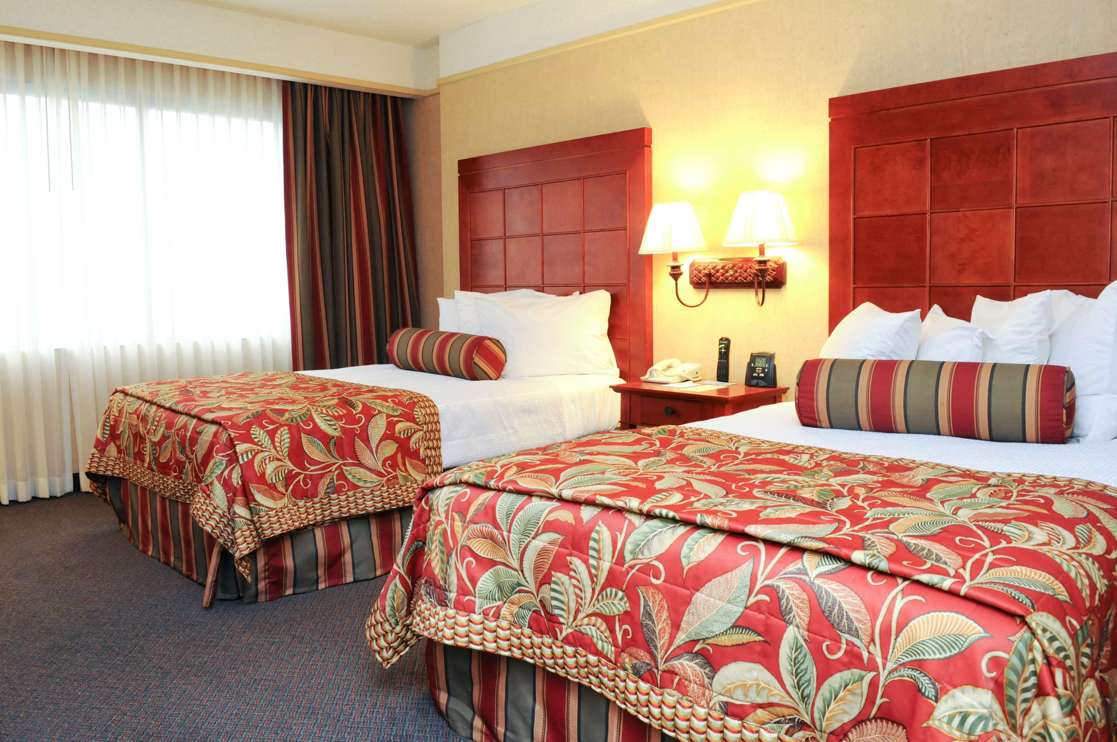 Habitación de hotel con dos camas, cada una con colcha de estampado floral, una mesita de noche con lámpara y una ventana con cortinas.