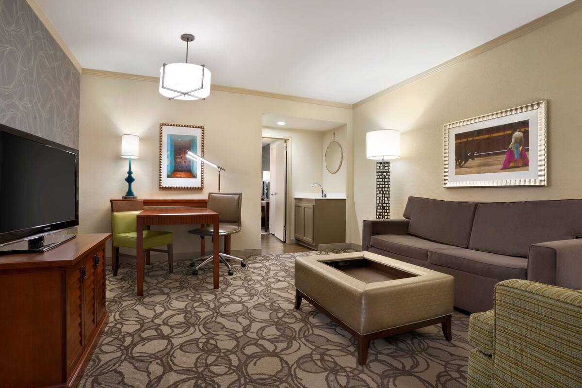 A hotel room with a flat-screen TV, a dining table with chairs, a sofa, a coffee table, and a patterned carpet.