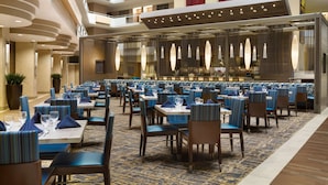 A spacious indoor dining area with multiple tables and chairs, a high ceiling with recessed lighting, and a view of the restaurant kitchen through a glass partition.