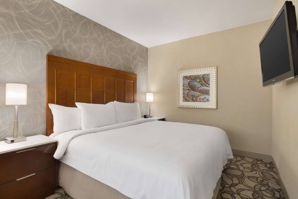 A hotel room with a large bed, two bedside tables with lamps, a flat-screen TV, and a decorative wall piece.