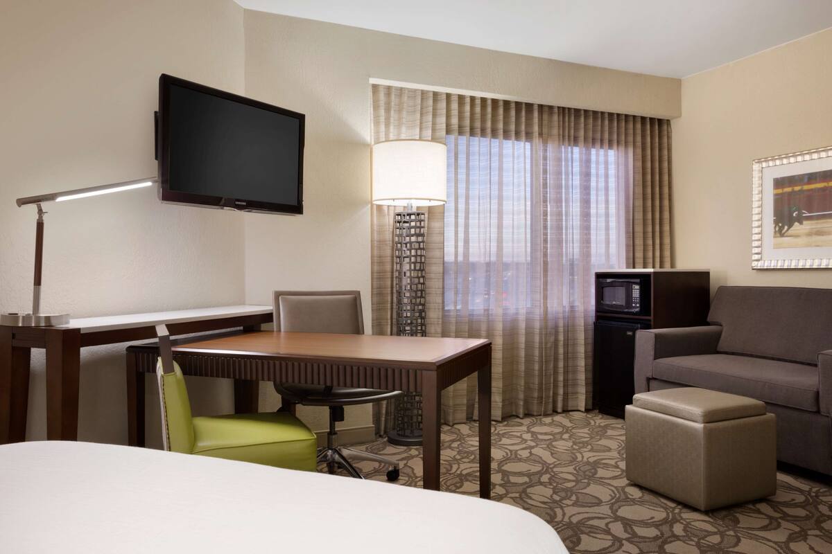 A hotel room with a flat-screen TV mounted on the wall, a desk with a chair, a sofa, a small ottoman, and a patterned carpet.