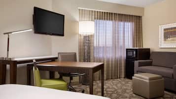 A hotel room with a flat-screen TV mounted on the wall, a desk with a chair, a sofa, a small ottoman, and a patterned carpet.