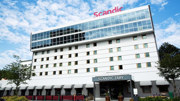Ein modernes Hotelgebäude mit blauem Himmel und dem Schriftzug "Scandic" auf der Spitze.