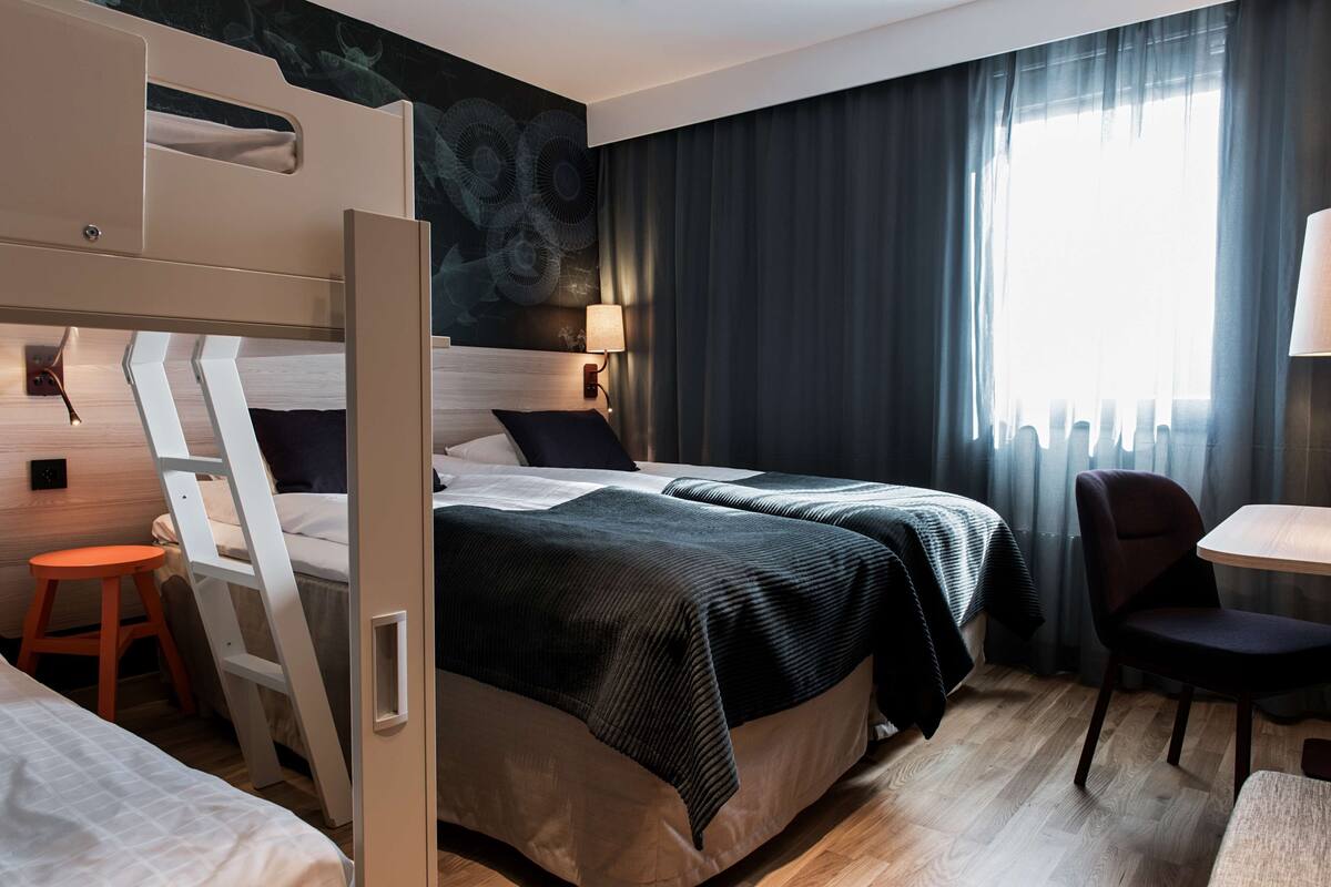 A hotel room with a bunk bed, a desk, a chair, a lamp, and a view of the outdoors.