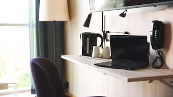 Ett hotellrum med ett skrivbord, en bärbar dator, en telefon, en kaffebryggare och en lampa.