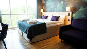 Ein Hotelzimmer mit einem großen Bett, einem Schreibtisch und einem Stuhl. Ein Fenster bietet Blick auf Bäume.