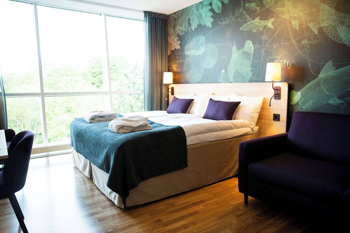 A hotel room with a large bed, a desk, and a chair. There is a window with a view of trees.