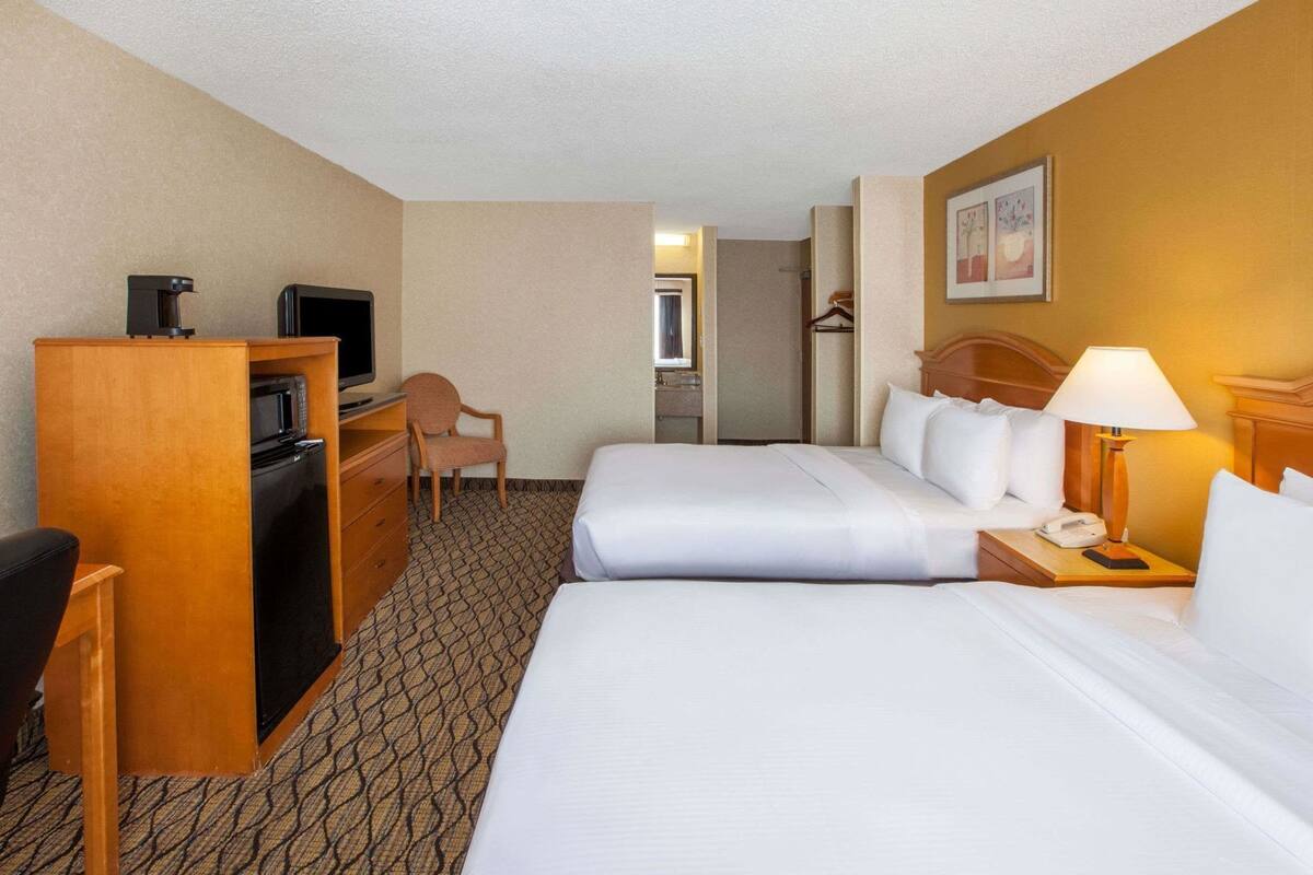 A hotel room with two beds, a desk with a TV, a chair, a lamp, and a small kitchenette.