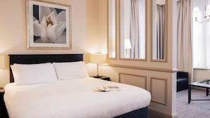 Une chambre d’hôtel avec un grand lit, une table de chevet, une lampe de lecture, un tableau encadré au mur, un miroir et une fenêtre avec des rideaux.