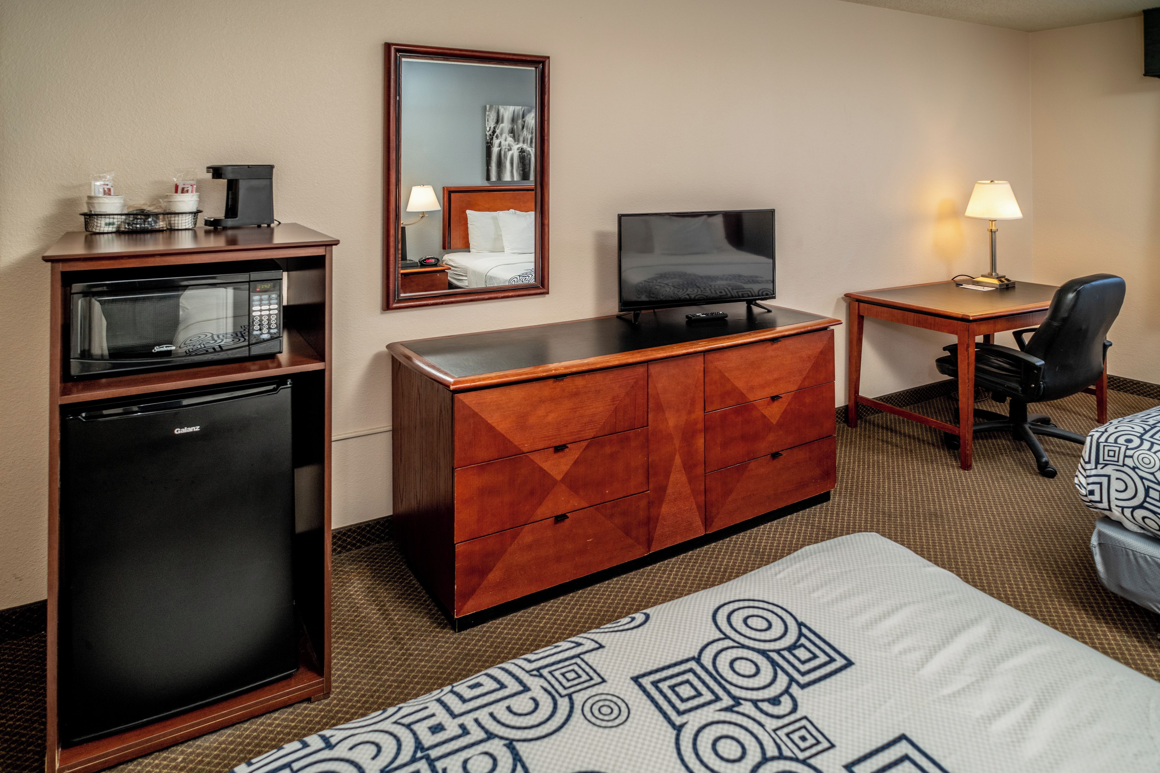 Een hotelkamer met een kitchenette, een bed, een bureau met stoel, een televisie en een spiegel.