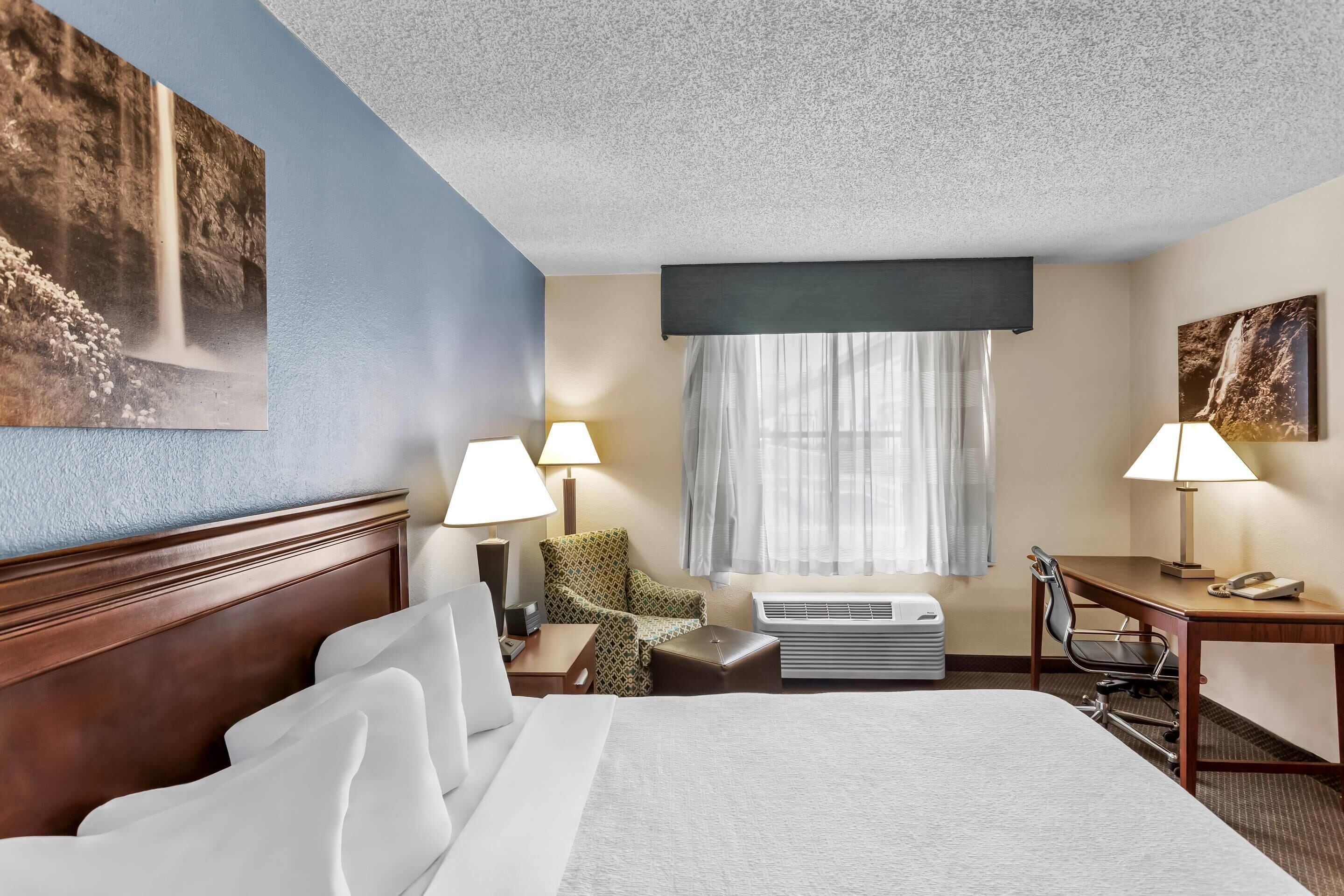 A hotel room with a bed, two chairs, a desk, an air conditioning unit, and a window with curtains.