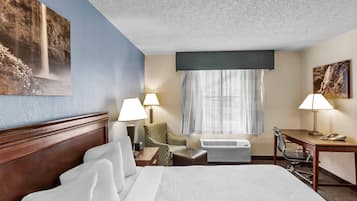 A hotel room with a bed, two chairs, a desk, an air conditioning unit, and a window with curtains.