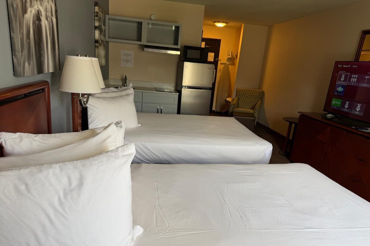 A hotel room with two beds, a desk, a chair, a TV, and a kitchenette.